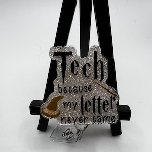 May include: A silver glitter badge holder with a black and gold design. The text reads "Tech because my letter never came".