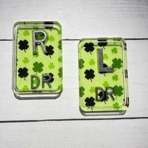May include: Two rectangular, clear acrylic pieces. One has a silver "R" and the text "DR" over a light green background with black shamrocks. The other has a silver "L" and "DR" over a similar background.