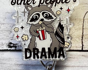 May include: A silver glitter badge holder with a cartoon raccoon eating popcorn. The badge holder has the text "Fueled by other people's drama" on it.