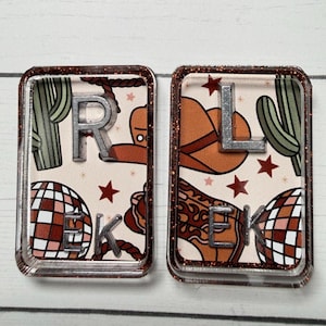 May include: Two rectangular resin keychains with a glittery brown border. One features the letters "R" and "E" and "K", the other "L" and "E" and "K". The background has a cowboy theme with cacti, a cowboy hat, and a disco ball.