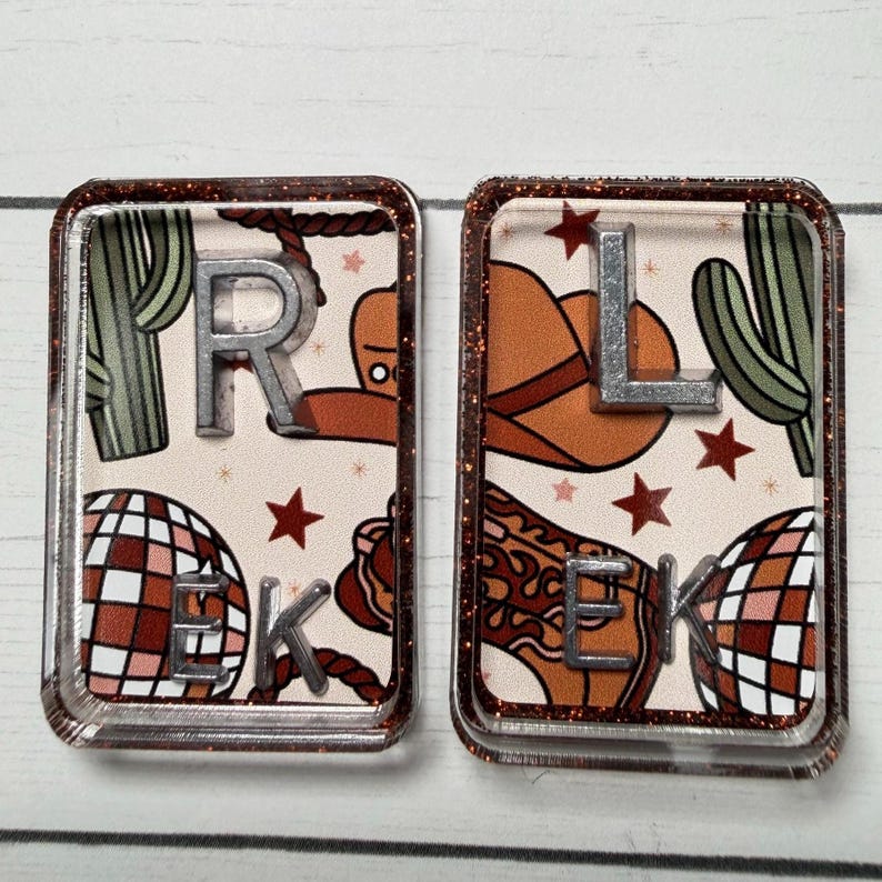 May include: Two rectangular resin keychains with a glittery brown border. One features the letters "R" and "E" and "K", the other "L" and "E" and "K". The background has a cowboy theme with cacti, a cowboy hat, and a disco ball.