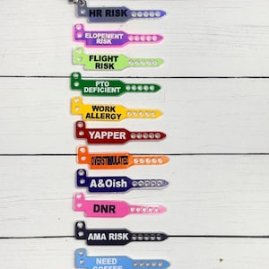 May include: A collection of eleven colorful acrylic keychains shaped like medical identification wristbands, each featuring black text including HR RISK, ELOPEMENT RISK, FLIGHT RISK, PTO DEFICIENT, WORK ALLERGY, YAPPER, OVERSTIMULATED, A&Oish, DNR, AMA RISK, and NEED COFFEE.