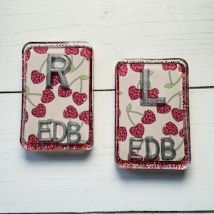May include: Two rectangular resin pieces with glitter edges. One has a silver "R" and the other a silver "L". Both have a cherry pattern background and the letters "EDB".