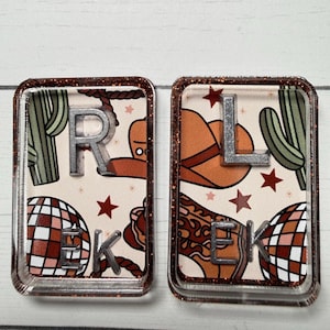 May include: Two rectangular keychains with a glittery brown border. One keychain features the letters "R", "E", and "K" with a cowboy theme, including a rope, cactus, and disco ball. The other keychain has the letters "L", "E", and "K" with a cowboy hat, cactus, and disco ball.