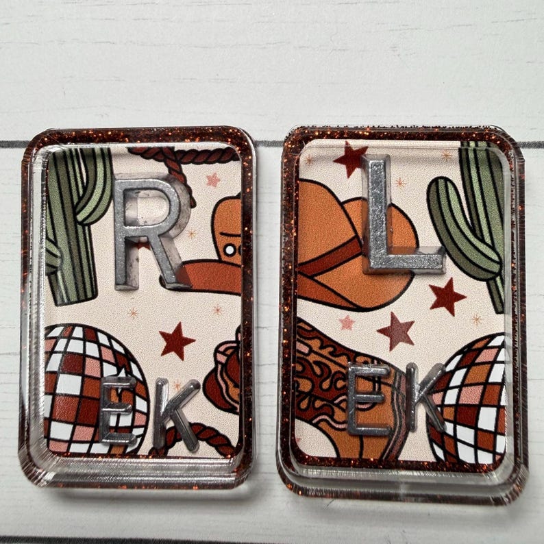 May include: Two rectangular keychains with a glittery brown border. One keychain features the letters "R", "E", and "K" with a cowboy theme, including a rope, cactus, and disco ball. The other keychain has the letters "L", "E", and "K" with a cowboy hat, cactus, and disco ball.