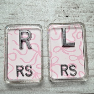 May include: Two rectangular, clear resin magnets. Each magnet has a light pink background with a bow pattern. One magnet has a silver "R" and "RS", the other has a silver "L" and "RS".