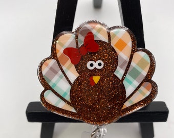 May include: A brown glitter turkey with a plaid patterned tail and a red bow. The turkey has white eyes and a yellow beak. The turkey is attached to a clear plastic retractable badge holder.