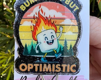 May include: A black and white glitter resin keychain with a retro sunset design and a cartoon marshmallow on fire. The text "Burnt Out But Optimistic" is written around the marshmallow. The keychain is attached to a silver key ring.