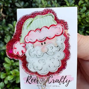 May include: A festive Santa Claus badge reel with a red glitter border. The Santa design features a white beard, pink cheeks, and a green and pink hat. The badge reel is attached to a white card with the text "ReeCrafty".