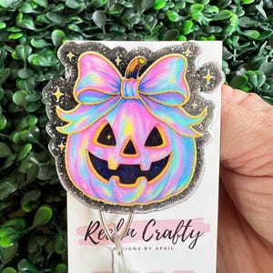 May include: A colorful Halloween-themed badge reel featuring a pastel pumpkin with a bow. The pumpkin has a smiling face and is outlined in gold. The badge reel has a glittery border and the text "Real n Crafty" is visible.
