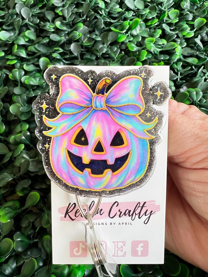 May include: A colorful Halloween-themed badge reel featuring a pastel pumpkin with a bow. The pumpkin has a smiling face and is outlined in gold. The badge reel has a glittery border and the text "Real n Crafty" is visible.