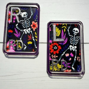 May include: Two rectangular, clear acrylic pins with a purple border. Each pin features a dancing skeleton design on a black background with colorful floral accents. One pin has the letter "R" and "RD", the other has "L" and "RD".