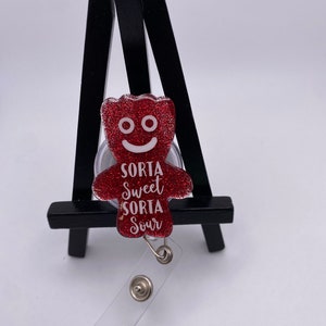 May include: Red glitter gummy bear charm with a white smiling face and the text "Sorta Sweet Sorta Sour" attached to a clear retractable badge holder.