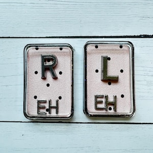 May include: Two rectangular, clear-framed accessories with a pink background. The left one has a silver "R" and the letters "EH" below, and the right one has a silver "L" and "EH". Each has three black dots.