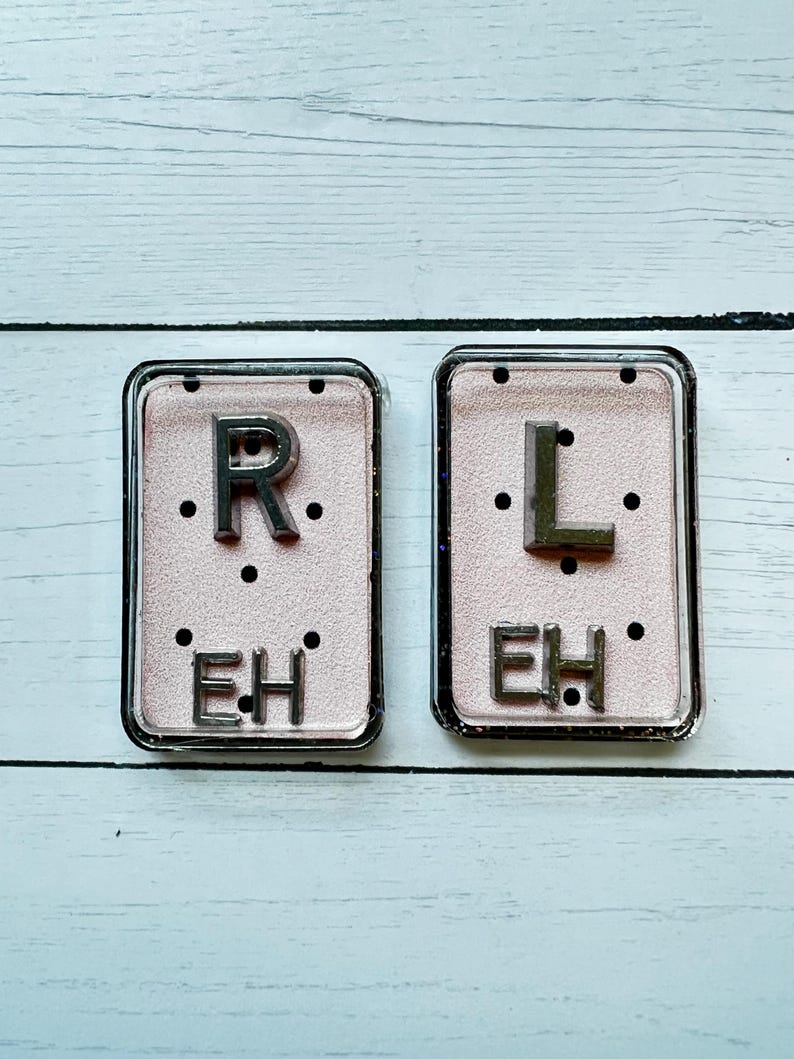 May include: Two rectangular, clear-framed accessories with a pink background. The left one has a silver "R" and the letters "EH" below, and the right one has a silver "L" and "EH". Each has three black dots.