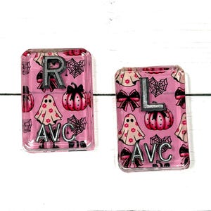 May include: Two rectangular acrylic charms featuring a pink background with a repeating pattern of ghosts, pumpkins, black bows, and spiderwebs. The left charm has the letters R and AVC, while the right charm has the letters L and AVC in silver.