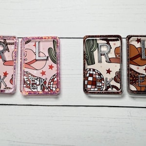 May include: Four rectangular resin keychains with a Western theme. Each keychain features a letter, a disco ball, a cowboy hat, and a cactus design. The keychains are in pink and brown tones with glitter accents.