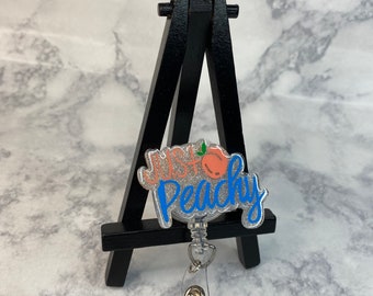 May include: A silver glitter badge holder with a blue and pink peach design and the text "Just Peachy".