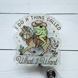 May include: A clear badge reel with a cartoon frog riding a horse, wearing a cowboy hat and saddle. The text reads "I do a thing called What I Want". The design includes cacti and glitter.