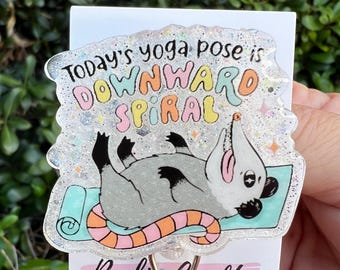 May include: A clear, glittery badge reel featuring a cartoon opossum in a yoga pose on a teal mat. The text reads "Today's yoga pose is downward spiral" in colorful letters. The reel has a metal clip and the words "Reelin' Crafty" are printed on the card.