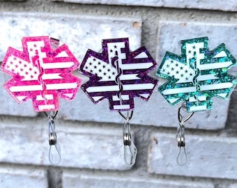 May include: Three glitter star of life badge reels with a white background and an American flag design. The reels are pink, purple, and teal.