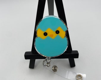 May include: A blue and yellow chick hatching from an egg, with glitter accents, attached to a clear plastic retractable badge holder.