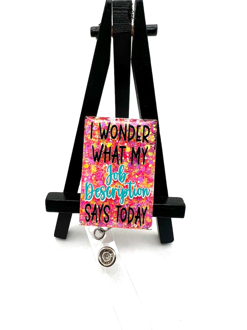 May include: A pink and yellow glitter badge holder with the text "I wonder what my job description says today".