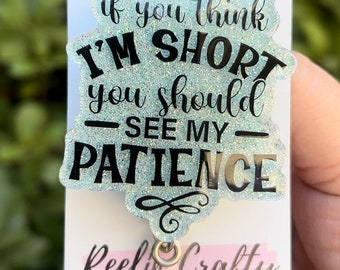 May include: A light blue glitter badge holder with the text "if you think I'M SHORT you should - SEE MY - PATIENCE" in black lettering. The badge holder is attached to a silver retractable badge reel. The badge holder is on a white card with the text "Reel'n Crafty DESIGNS BY APRIL" in black lettering.