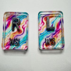 May include: Two rectangular resin pieces with a colorful, swirled background of pink, blue, and purple. One piece has a silver "R" and "WS" text, the other has a silver "L" and "WS" text. The pieces are likely for a craft project.