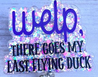 Puede incluir: Un portatarjetas con un diseño brillante e iridiscente. El texto de la imagen dice "Welp, There Goes My Last Flying Duck" en letras moradas y negras. El carrete tiene un clip de metal y una correa de plástico transparente.