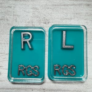 May include: Two rectangular teal-colored objects with silver lettering. The left object has the letter "R" and the letters "RGS" below it. The right object has the letter "L" and the letters "RGS" below it.