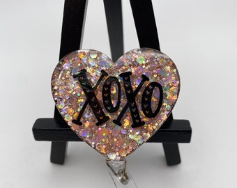 May include: A pink glitter heart-shaped badge holder with a black "XOXO" design and a clear retractable badge reel.