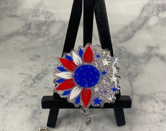 May include: A red, white, and blue glitter sunflower badge reel with a clear plastic clip. The sunflower has a blue center with white petals and red accents.