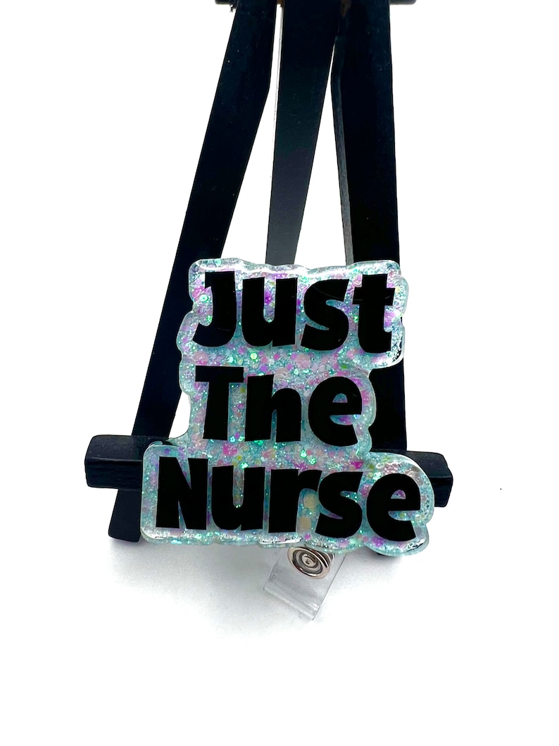 May include: A light blue and iridescent glitter badge holder with a black text overlay that reads "Just The Nurse".
