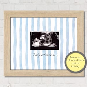 May include: A light wood frame containing a white mat with vertical light blue watercolor stripes. Centered is a black and white ultrasound image above the text Baby Henderson in a dark blue script font. A circular badge reads More mat colors and frame options in lising.