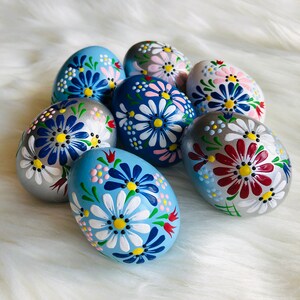 Può includere: Otto uova di Pasqua dipinte a mano con motivi floreali. Le uova sono dipinte in tonalità di blu, argento, rosso, bianco e rosa. I motivi presentano fiori, foglie e punti.