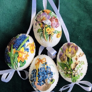 Könnte beinhalten: Vier weiße Ostereier mit Blumenmustern. Jedes Ei hat ein anderes Blumenmuster, darunter Tulpen, Hyazinthen und Krokusse. Die Eier sind mit weißen Bändern verziert.