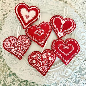 May include: Six red heart-shaped decorations with white embroidered patterns and scalloped edges. Each has a white ribbon for hanging. The hearts are arranged on a white, ornate decorative plate.