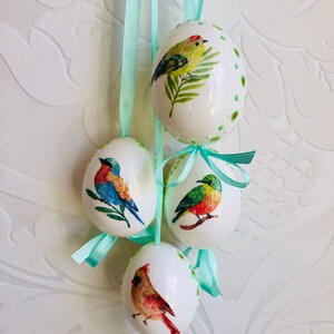 Könnte beinhalten: Vier weiße Ostereier mit grünen Schleifen zum Aufhängen. Jedes Ei ist mit einer anderen Vogel-Illustration verziert. Die Vögel sind ein Kardinal, ein Blauvogel, ein grüner Vogel und ein gelber Vogel.
