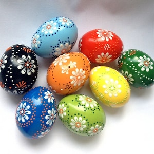 Könnte beinhalten: Eine Sammlung handbemalter Ostereier in verschiedenen Farben, darunter Blau, Rot, Grün, Orange und Gelb. Jedes Ei ist mit einem weißen Blumenmuster und kleinen Punkten verziert. Die Eier sind auf einer weißen Oberfläche angeordnet.