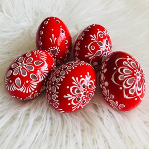 Könnte beinhalten: Fünf rote Ostereier, die mit weißen Blumenmustern und filigranen Ausschnitten verziert sind. Die Eier liegen auf einer weißen, flauschigen Oberfläche.