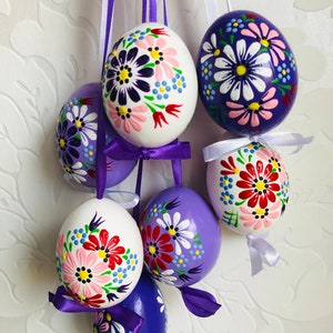 Può includere: Sei uova di Pasqua dipinte a mano con motivi floreali. Le uova sono bianche e viola con fiori rosa, blu, gialli e verdi. Le uova sono appese a nastri bianchi e viola.