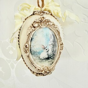 Op de afbeelding: Crèmekleurig paasei-ornament met een ingelijste afbeelding van een wit konijn in een tuinlandschap. Het ei heeft een decoratieve rand en is afgewerkt met een gele lint en een touwlus om op te hangen.