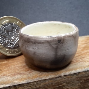 May include: A small, handmade ceramic bowl with a light brown exterior and a cream-colored interior. The bowl is next to a United Kingdom one-pound coin. The bowl is approximately 5 cm in diameter and 4 cm tall.