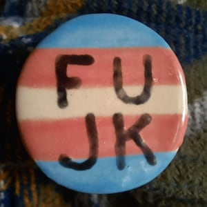 May include: A circular badge with a transgender pride flag design. The badge has a blue top and bottom, a white stripe in the middle, and a pink stripe. The letters "FU JK" are written in black across the badge.