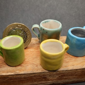 Pode incluir: Quatro canecas de cerâmica em miniatura em diferentes cores: verde, amarelo, azul e verde com manchas marrons. As canecas estão dispostas sobre uma superfície de madeira.