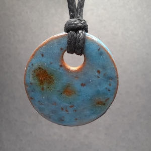May include: A round ceramic pendant with a blue glaze and brown speckles. The pendant has a hole in the centre and is attached to a black cord.