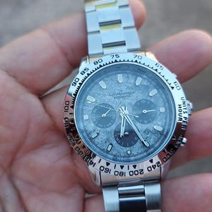 May include: A silver-toned wristwatch with a textured gray face and a matching metal band. The watch features three sub-dials and a bezel with numerical markings. The watch is held in a hand.