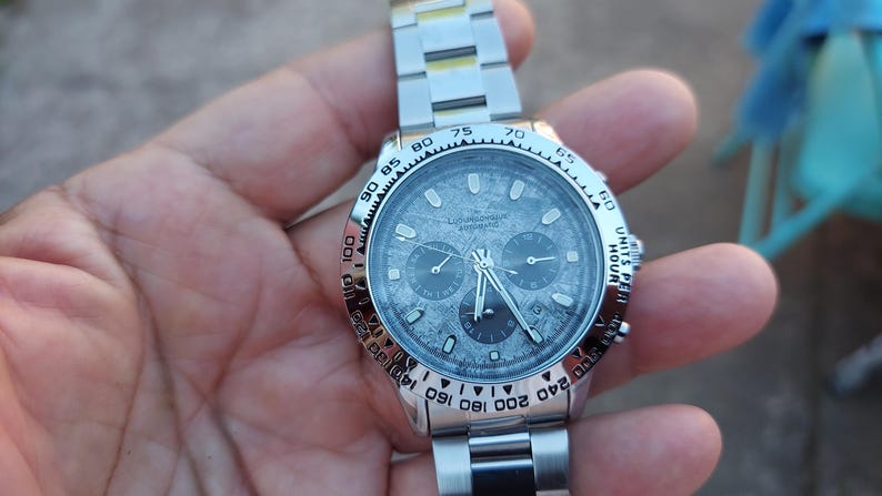 May include: A silver-toned wristwatch with a textured gray face and a matching metal band. The watch features three sub-dials and a bezel with numerical markings. The watch is held in a hand.