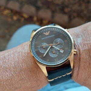 May include: Gold-toned wristwatch with a brown face and black leather band. The watch features multiple subdials and the brand name "Emporio Armani" is visible on the face. The watch is secured on a wrist.